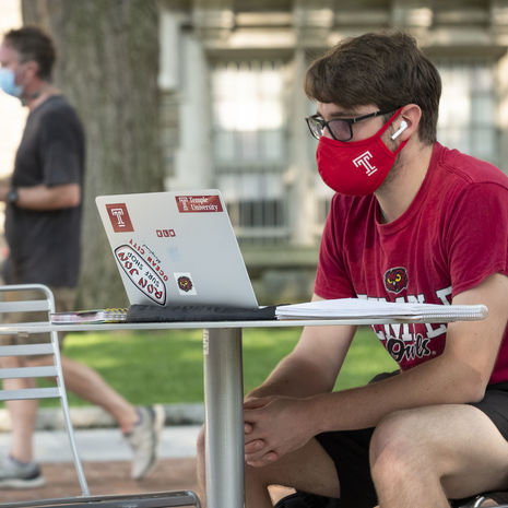 A student sitting outside on campus wearing a mask while using their computer. 