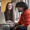 Two students smiling and looking at a laptop on a table.