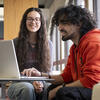 Two students smiling and looking at a laptop on a table.